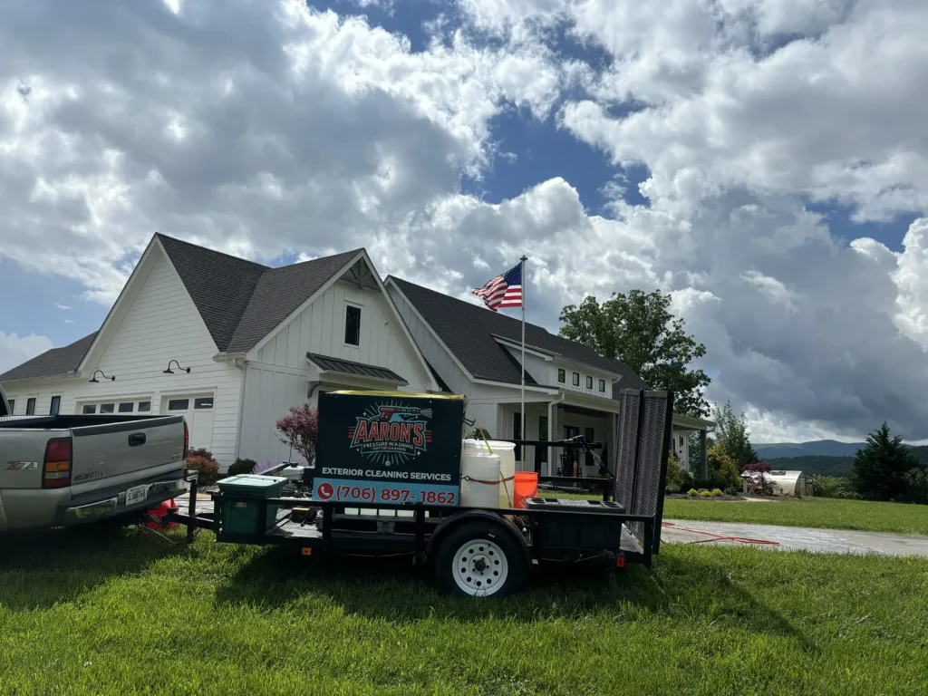 A service trailer for "Aaron's Exterior Cleaning Services" parked in the yard of a white modern farmhouse with a dark roof and an American flag flying, under a cloudy sky